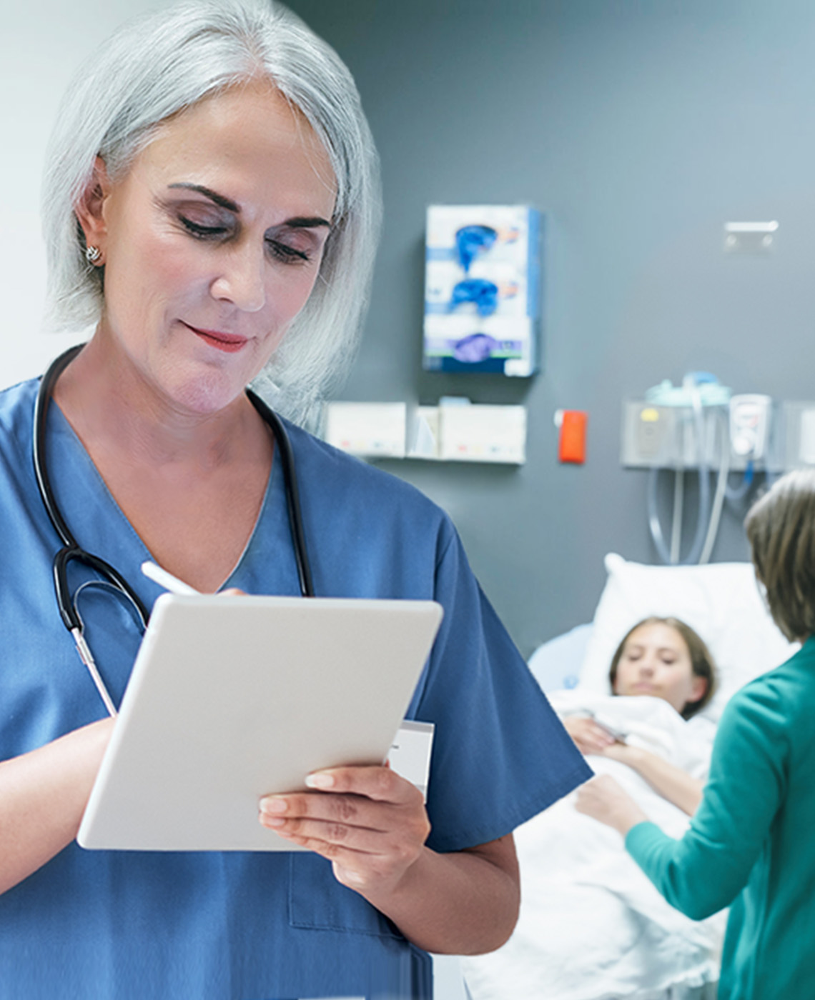 A nurse takes notes on a tablet with a stylus. Behind her, a patient lies in a hospital bed with a visitor at her bedside.